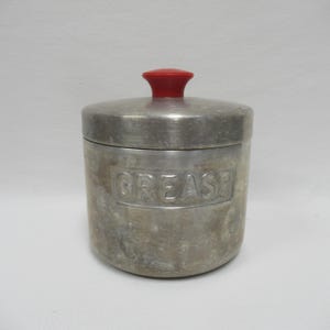 May include: A vintage metal grease container with a red knob on the lid. The word "GREASE" is embossed on the front of the container. The container has a cylindrical shape and a silver finish.