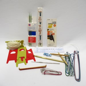 May include: Assortment of knitting supplies including needles, yarn, a yarn threader, stitch markers, and a gauge ruler. The needles are packaged in clear plastic with blue and white labels. The yarn is white and wound on a cardboard spool. The yarn threader is packaged in a white cardboard box with a black and white illustration of a yarn threader. The stitch markers are red, green, and yellow. The gauge ruler is white with black markings.