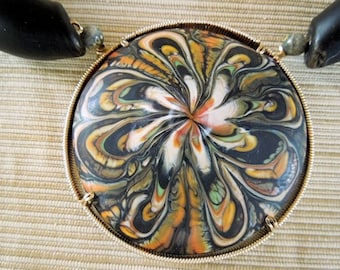 May include: A round, gold-rimmed pendant with a swirling black, white, orange, and green pattern. The pendant is attached to a black cord.