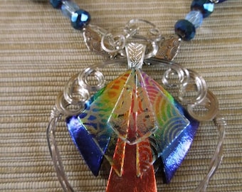 May include: A silver wire heart-shaped pendant with a colorful, iridescent, geometric glass piece hanging from it. The glass piece is made up of multiple layers of glass, each with a different color and pattern. The pendant is attached to a silver chain with blue and clear glass beads.