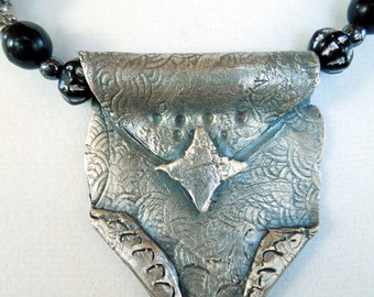 May include: A silver necklace with a textured, patterned pendant shaped like a small pouch. The pendant has a star-shaped design in the center.