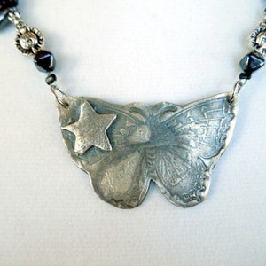 May include: A silver necklace with a butterfly pendant featuring a star. The butterfly is detailed with etched lines and the star is in the center of the butterfly's wings. The necklace is made with black beads and silver accents.