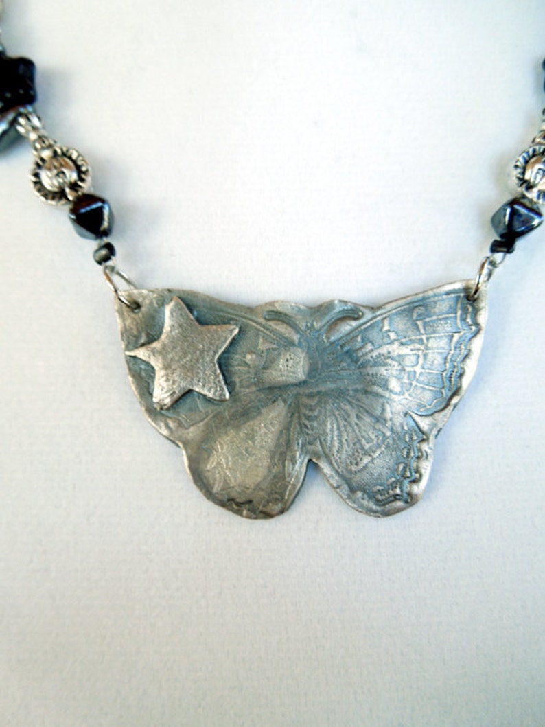 May include: A silver necklace with a butterfly pendant featuring a star. The butterfly is detailed with etched lines and the star is in the center of the butterfly's wings. The necklace is made with black beads and silver accents.