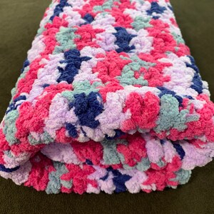 May include: A soft, crocheted blanket in shades of pink, purple, blue, and green. The blanket is made with a chunky yarn and has a fluffy texture.