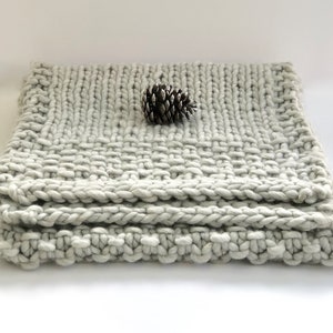 May include: A soft, gray knitted throw blanket with a textured weave. The blanket is folded in thirds and a small pine cone is resting on top.