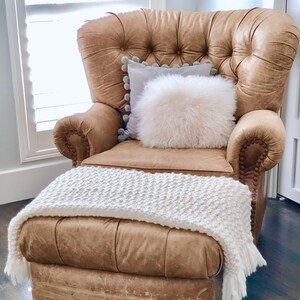 May include: A brown leather armchair with a white faux fur pillow and a white knitted throw blanket. The chair has a tufted back and a button-tufted design.