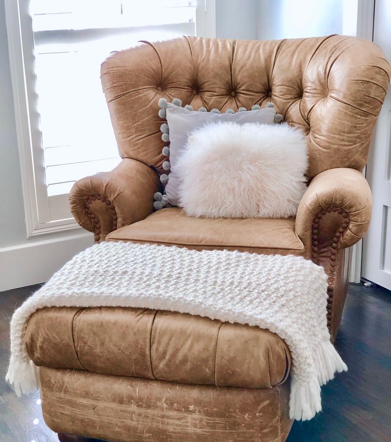 May include: A brown leather armchair with a white faux fur pillow and a white knitted throw blanket. The chair has a tufted back and a button-tufted design.