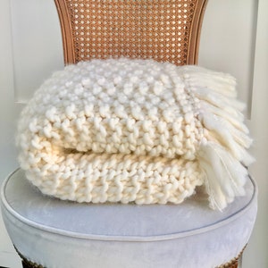 May include: A white knitted throw blanket with a fringe on the edges, folded and placed on a light gray upholstered chair with a wooden frame.