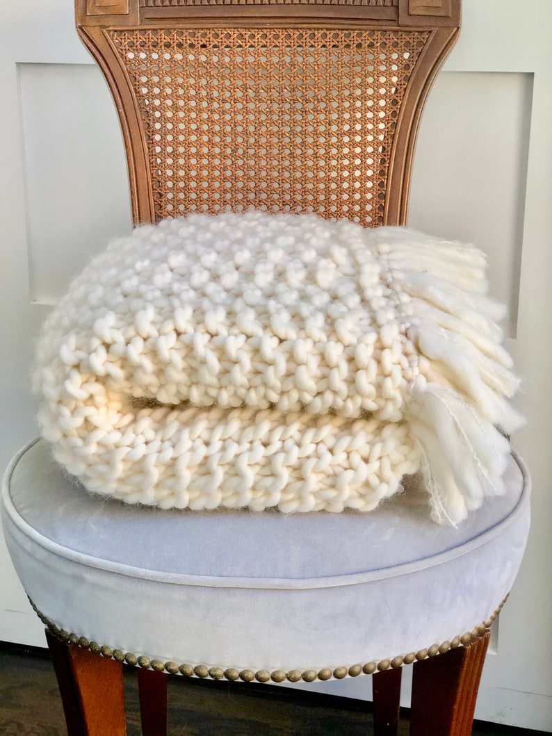 May include: A white knitted throw blanket with a fringe on the edges, folded and placed on a light gray upholstered chair with a wooden frame.