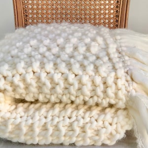 May include: A white chunky knit throw blanket with a fringe border. The blanket is folded and sitting on a wicker chair.