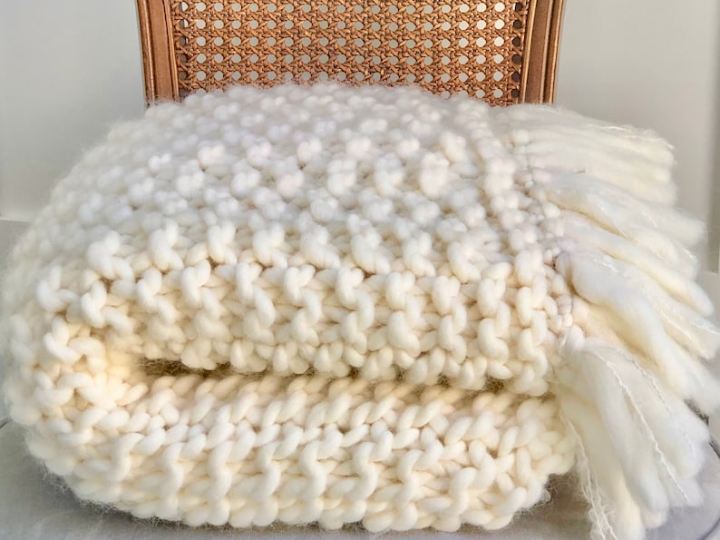 May include: A white chunky knit throw blanket with a fringe border. The blanket is folded and sitting on a wicker chair.