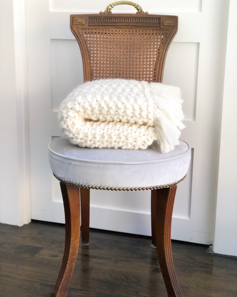 May include: A white knitted throw blanket with fringe is folded and placed on a light gray upholstered chair with a wooden frame and cane back.