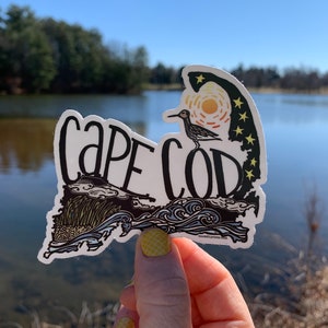 Op de afbeelding: Een witte sticker met een zwarte omtrek van Cape Cod, Massachusetts. De sticker heeft een gestileerde vogel, golven en een zonsondergang met sterren. De tekst "Cape Cod" staat in het zwart bovenaan de sticker. De sticker wordt in een hand gehouden tegen een achtergrond van een meer en bomen.