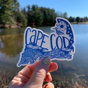 Op de afbeelding: Een blauwe en witte sticker met de woorden "Cape Cod" in een gestileerd lettertype. De sticker heeft de vorm van de staat Massachusetts en bevat een vogel en sterren.