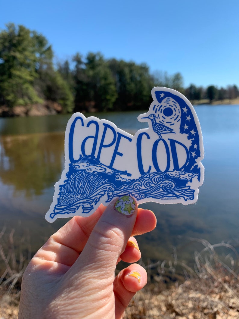 Op de afbeelding: Een blauwe en witte sticker met de woorden "Cape Cod" in een gestileerd lettertype. De sticker heeft de vorm van de staat Massachusetts en bevat een vogel en sterren.
