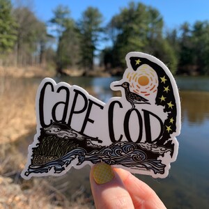 Op de afbeelding: Een witte sticker met een zwarte omtrek van de staat Massachusetts, met de woorden "Cape Cod" in zwarte cursieve letters. De sticker heeft een gestileerde illustratie van een vogel, golven en een zon met sterren. De sticker wordt in een hand gehouden tegen een wazige achtergrond van bomen en water.
