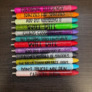 May include: A collection of glitter pens in various colors, each with a different phrase in black lettering. The pens have different colored tips and are arranged vertically against a dark background. The phrases include "IDAZODONE DEFICIENCY" and "DON'T TOUCH MV DEN."