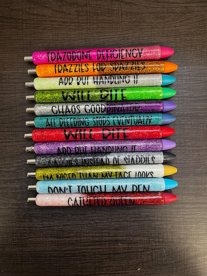May include: A collection of glitter pens in various colors, each with a different phrase in black lettering. The pens have different colored tips and are arranged vertically against a dark background. The phrases include "IDAZODONE DEFICIENCY" and "DON'T TOUCH MV DEN."