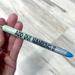 May include: A glittery pen with a gradient design, transitioning from light green to silver. The pen features the text "ADD OUT HANDLING IT" in black, with a blue tip. The pen is held in a hand.