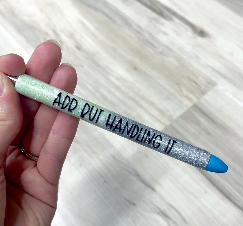 May include: A glittery pen with a gradient design, transitioning from light green to silver. The pen features the text "ADD OUT HANDLING IT" in black, with a blue tip. The pen is held in a hand.