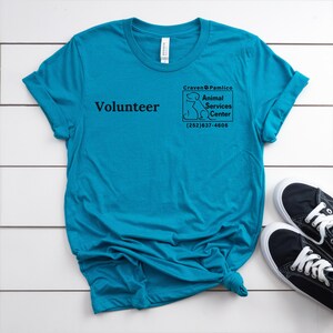May include: A turquoise blue t-shirt with the text "Volunteer" and a logo for the Craven Pamlico Animal Services Center. The logo includes a silhouette of a dog and the phone number (252) 637-4606.