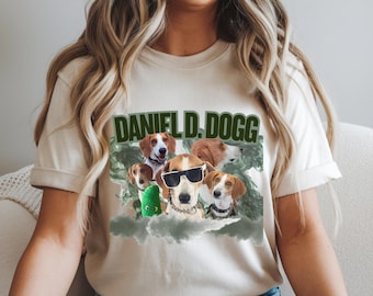 May include: Beige t-shirt featuring a graphic of several dogs, one wearing sunglasses, with the text "DANIEL D. DOGG" in green. The dogs are set against a cloudy, green-tinted background.