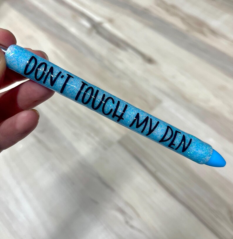 May include: A blue glitter pen with the text "DON'T TOUCH MY DEN" in black. The pen has a blue tip and a silver metal loop at the top. The pen is held in a hand.