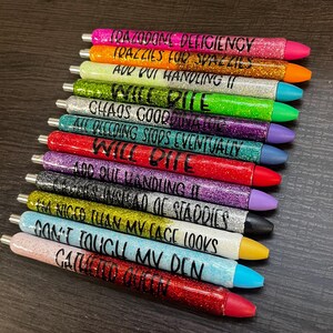 May include: A collection of glitter pens in various colors, each with a different phrase in black lettering. The pens have a metallic silver tip and a colored tip. The phrases include "TRAZZIES FOR SPAZZIES" and "DON'T TOUCH MY DEN."
