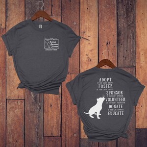 May include: Gray t-shirt with a white graphic on the front that says "Craven Pamlico Animal Services Center (252)637-4606". The back of the shirt has a white graphic of a dog with the text "Adopt if you can't adopt, Foster if you can't foster, Sponsor if you can't sponsor, Volunteer if you can't volunteer, Donate if you can't donate, Educate".