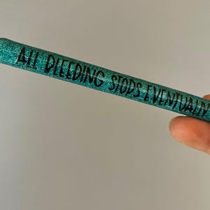 May include: A teal glitter pen with the phrase "ALL BLEEDING STOPS EVENTUALLY" in black lettering. The pen is held in a hand against a light gray background. The pen has a silver metal tip.