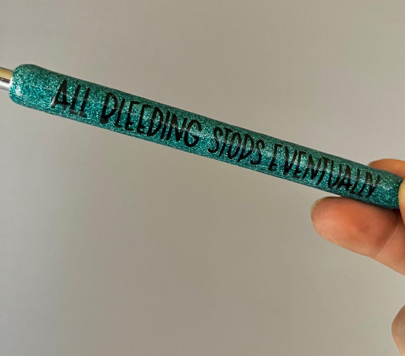 May include: A teal glitter pen with the phrase "ALL BLEEDING STOPS EVENTUALLY" in black lettering. The pen is held in a hand against a light gray background. The pen has a silver metal tip.