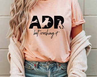 May include: A pink t-shirt with black text that reads "ADR but crushing it". The text is made up of silhouettes of dogs performing agility tasks.
