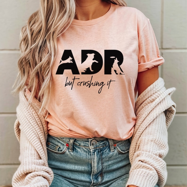 May include: A pink t-shirt with black text that reads "ADR but crushing it". The text is made up of silhouettes of dogs performing agility tasks.