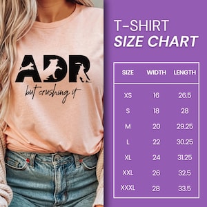 May include: A pink t-shirt with a black graphic that says "ADR but crushing it" and features three dog silhouettes. A size chart is shown on the right side of the image with the following sizes: XS, S, M, L, XL, XXL, XXXL. The chart lists the width and length of each size in inches.