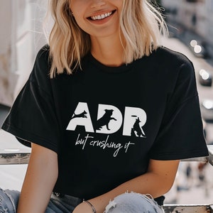 May include: A black t-shirt with a white graphic design that says "ADR but crushing it" with silhouettes of dogs playing fetch.