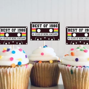 May include: Three cupcakes with white frosting and colorful sprinkles, each topped with a rectangular paper topper shaped like a cassette tape. The toppers feature the text BEST OF 1986 and 40 YEARS OF BEING AWESOME with rainbow accents.