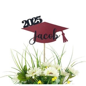 May include: A maroon graduation cap cake topper featuring the black text 2025 and the name Jacob in black script. The topper is attached to a thin wooden stick and is positioned above a bouquet of white flowers with green leaves and grass.