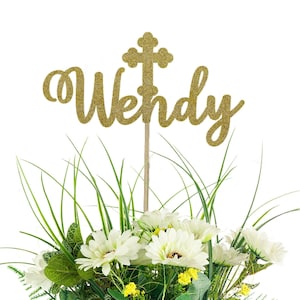 May include: A gold glitter cake topper featuring the name Wendy in cursive script with a cross symbol replacing the letter t. The topper is attached to a thin wooden stick and is positioned above a bouquet of white flowers and green foliage.