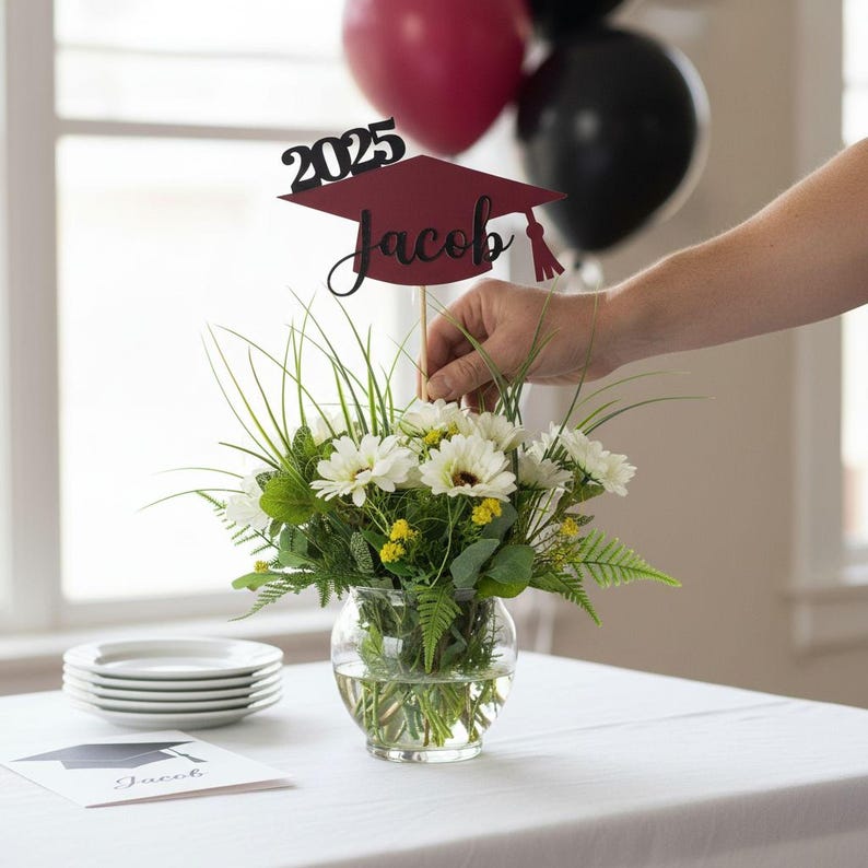 A person is arranging a bouquet of white flowers in a glass vase on a table, with a graduation cap-shaped topper and balloons in the background.