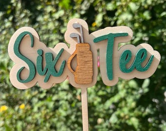 May include: A wooden cake topper featuring the text Six Tee in dark green script, with a brown wooden golf bag icon replacing the letter I. The design is mounted on a light wood base and includes a thin wooden stake for insertion into a cake.