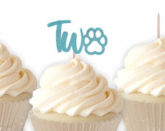 May include: Three cupcakes with white frosting in silver liners. The center cupcake features a light blue glittery cake topper with the word Two and a paw print icon attached to a thin wooden pick, while the others have plain wooden picks.