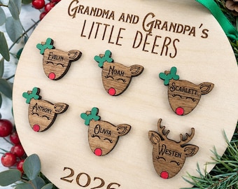 May include: A round wooden Christmas ornament featuring the engraved text "GRANDMA AND GRANDPA'S LITTLE DEERS" and "2026". It includes six small wooden deer head cutouts with red noses, green bows, and the names Evelyn, Noah, Scarlett, Anthony, Olivia, and Westen.