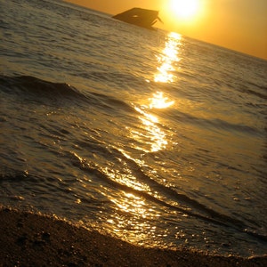 May include: A close-up of a sandy beach with the sun setting over the ocean. The water is reflecting the golden light of the sun, creating a shimmering effect.