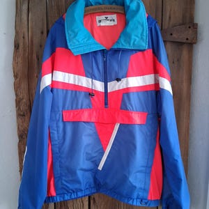 May include: A vintage blue, red, and white anorak jacket with a high collar and a half-zip closure. The jacket features a color-blocked design with white stripes and the text "OF EVER". The jacket is hanging on a wooden surface.