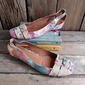 Puede incluir: Un par de zapatos planos coloridos con un estampado floral y de piel de serpiente. Los zapatos tienen un diseño de tiras en la parte trasera y están sobre una superficie de madera. Los zapatos son una mezcla de rosa, azul y amarillo.