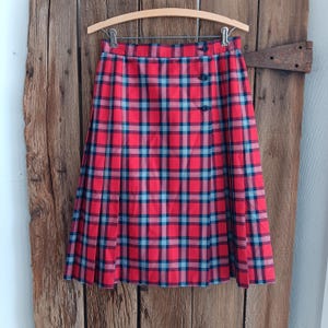Pode incluir: Uma saia de xadrez vermelha, preta e azul com fecho de botão. A saia é plissada e tem um comprimento até ao joelho.