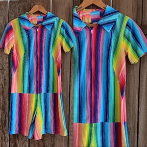 May include: A vibrant rainbow striped romper with a collared neckline and short sleeves. The colourful garment features a vertical stripe pattern in shades of red, orange, yellow, green, blue, and purple. It has a zip closure and is displayed on a wooden hanger.
