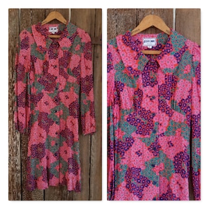 Könnte beinhalten: Ein langärmeliges Kleid mit floralem Druck in Rosa, Grün und Blau mit einem Kragen und Knopfverschluss. Das Kleid hängt an einem Holzbügel. Das Markenetikett lautet "Plain Jane".