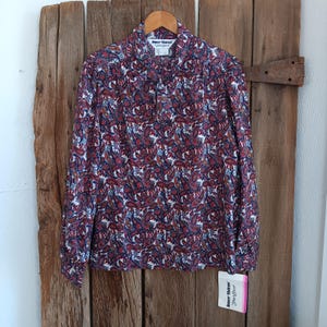 Puede incluir: Una blusa de manga larga con estampado de cachemira en rojo, azul y blanco cuelga de una percha de madera. La blusa tiene cuello abotonado y puños. Una etiqueta con el nombre de la marca está sujeta en la parte inferior. La blusa se exhibe sobre un fondo de madera rústica.