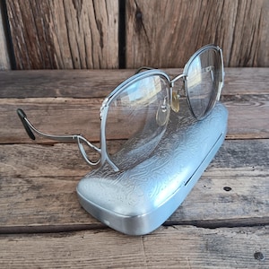 May include: Silver-framed spectacles with clear lenses sit on a silver case with an embossed floral pattern. The spectacles have a retro design with a thin silver frame and dark earpieces. The case is rectangular.